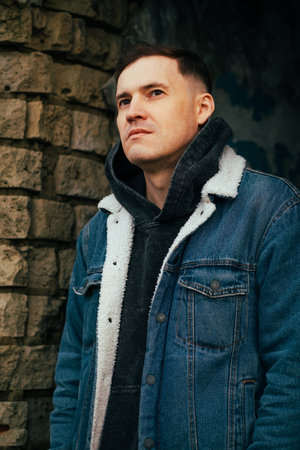 A man with short hair poses confidently against a weathered stone wall He wears a denim jacket over...