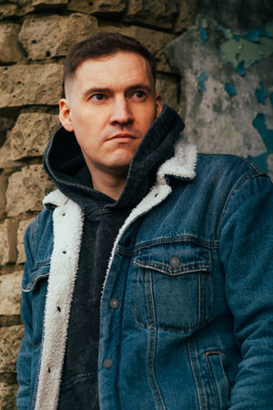 Man in Denim Jacket Stands Against Textured Stone Wall in Urban Setting During Late Afternoon Light