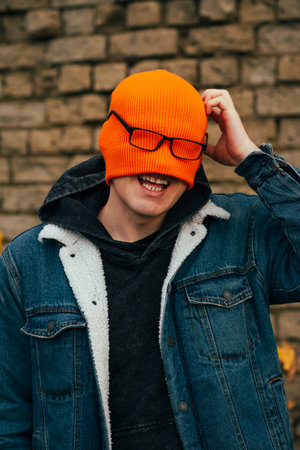 A young man is smiling while playfully adjusting his bright orange beanie which covers his face He stands outdoors next to a textured brick wall surrounded by falling autumn leavesの写真素材