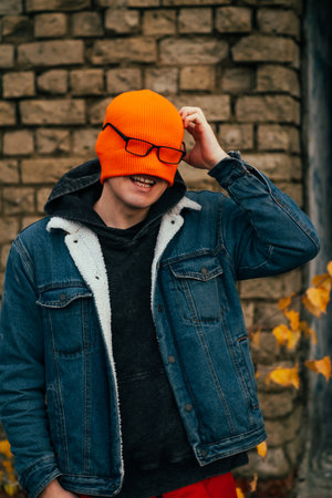A young man is smiling while playfully adjusting his bright orange beanie which covers his face He stands outdoors next to a textured brick wall surrounded by falling autumn leavesの写真素材