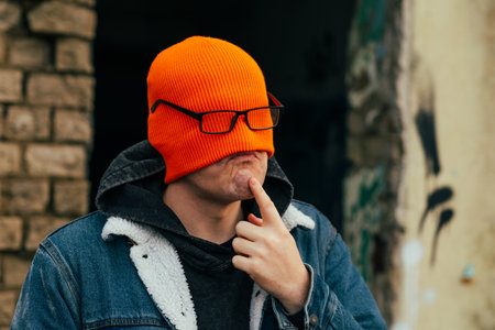 Unique Urban Portrait of a Man With Orange Beanie and Glasses Expressing Curiosity in a City Settingの写真素材