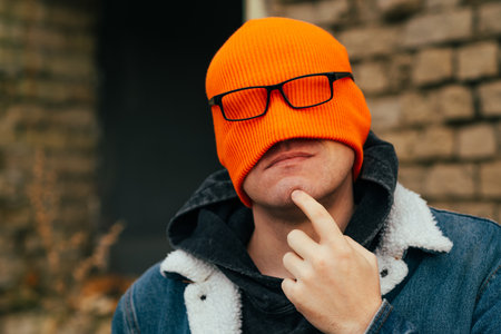Unique Urban Portrait of a Man With Orange Beanie and Glasses Expressing Curiosity in a City Settingの写真素材