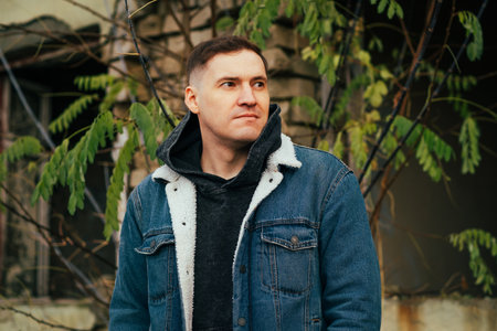 A man stands outside dressed in a denim jacket and hood He is surrounded by plants looking...
