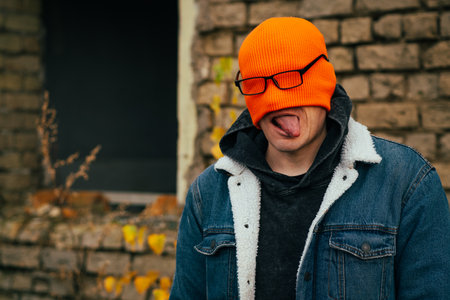 A young man stands in front of a brick wall wearing an orange beanie and glasses playfully sticking out his tongue The urban environment is punctuated by fallen leaves and a windowの写真素材