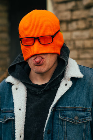 A young man stands in front of a brick wall wearing an orange beanie and glasses playfully sticking out his tongue. The urban environment is punctuated by fallen leaves and a window.の写真素材