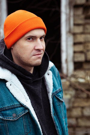 Young Man in Denim Jacket and Orange Beanie Stands Outside Abandoned Building During Autumn Afternoon.の写真素材