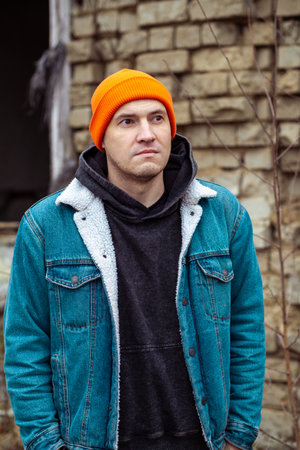 A young man wearing a denim jacket and orange beanie poses near an old stone building The area shows signs of neglect with overgrown plants It is a cool autumn afternoon setting a reflective mood.の写真素材