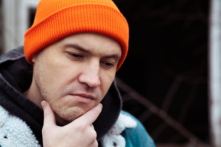 A young man wearing a denim jacket and orange beanie poses near an old stone building The area shows signs of neglect with overgrown plants It is a cool autumn afternoon setting a reflective mood.の写真素材