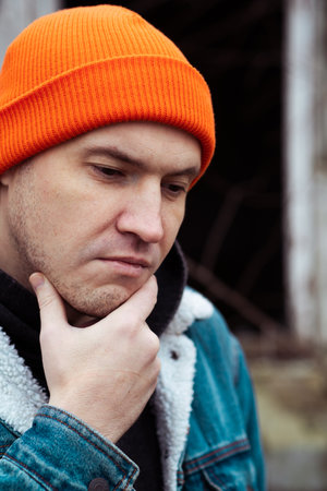 A young man wearing a denim jacket and orange beanie poses near an old stone building The area shows...
