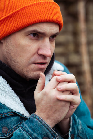 A man with an orange beanie and denim jacket is outside in a cold environment He is holding his hands together and gazing ahead suggesting deep thought or concernの写真素材