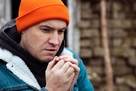 A man with an orange beanie and denim jacket is outside in a cold environment He is holding his hands together and gazing ahead suggesting deep thought or concernの写真素材