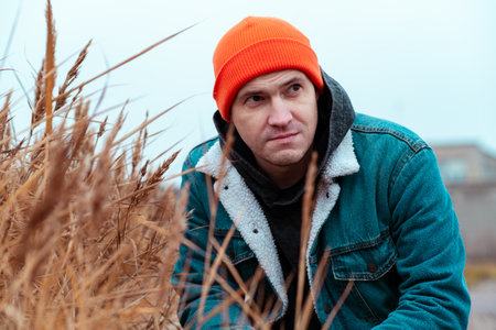 A man wearing an orange beanie and a blue jacket crouches among tall grass in a desolate abandoned location The cloudy sky adds to the gloomy atmosphereの写真素材
