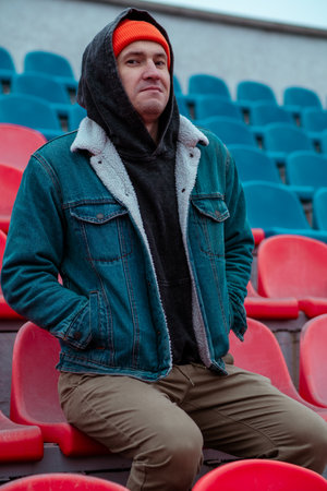 Man in a Red Beanie Sits on Empty Stadium Seats During a Chilly Day in a Quiet Outdoor Arenaの写真素材