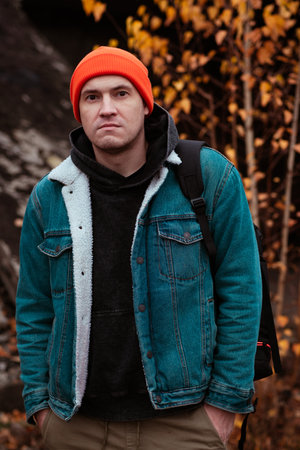 A young man stands outside wearing an orange beanie and denim jacket surrounded by colorful autumn leaves He looks thoughtful against a weathered backdrop of an urban environmentの写真素材