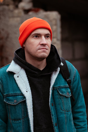 A young man stands outside wearing an orange beanie and denim jacket. He looks thoughtful against a weathered backdrop of an urban environment.の写真素材