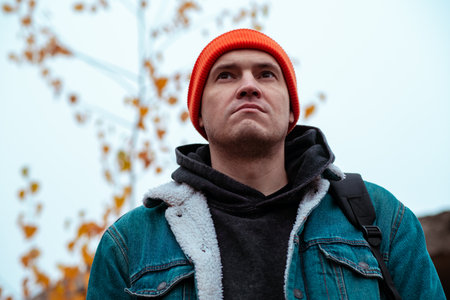 A young man stands outdoors on a cool autumn day wearing a colorful beanie and a denim jacket He gazes thoughtfully at the cloudy sky surrounded by fall foliageの写真素材