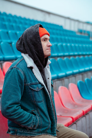 A person wearing a red beanie and denim jacket sits thoughtfully among empty red and blue stadium seats. The cloudy sky suggests a cool crisp day creating a reflective atmosphere.の写真素材