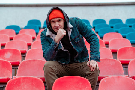 A person wearing a red beanie and denim jacket sits thoughtfully among empty red and blue stadium...
