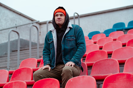 Man in a Red Beanie Sits on Empty Stadium Seats During a Chilly Day in a Quiet Outdoor Arenaの写真素材