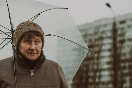 A woman stands outdoors on a rainy day, thoughtfully holding a transparent umbrella. She appears reflective amidst a backdrop of gray buildingsの写真素材