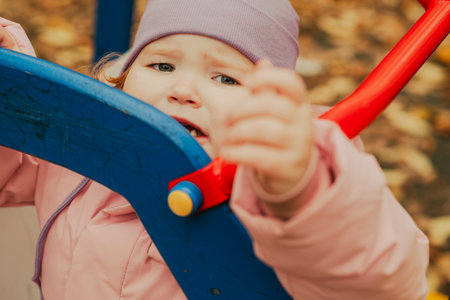 A young child, dressed warmly in a pink jacket and purple hat, appears to be having a mixed experience on a playful structure. Leaves scatter the ground, indicating autumn.の写真素材