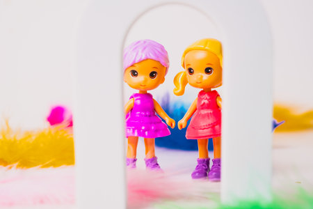 Two cheerful toy dolls stand hand in hand inside a small white arch, surrounded by colorful feathers. The dolls are dressed in bright outfits, creating a fun and playful scene.の写真素材