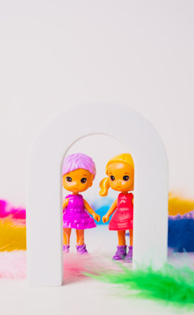Two cheerful toy dolls stand hand in hand inside a small white arch, surrounded by colorful feathers. The dolls are dressed in bright outfits, creating a fun and playful scene.の写真素材