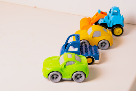 Four colorful toy vehicles are arranged in a row on a light wooden surface. The toys include a yellow truck, a blue car, a green vehicle, and an orange construction machine.の写真素材