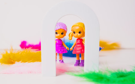 Two cheerful toy dolls stand hand in hand inside a small white arch, surrounded by colorful feathers. The dolls are dressed in bright outfits, creating a fun and playful scene.の写真素材