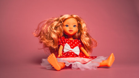 A doll with curly blonde hair sits on a pink background dressed in a red dress with white polka...