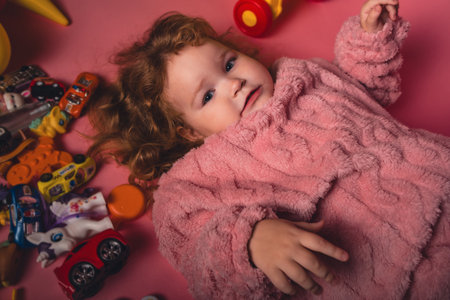 A young child lies on a pink floor surrounded by colorful toys. The child smiles widely while wearing a cozy sweater. There are many toy cars and figures scattered around.の写真素材