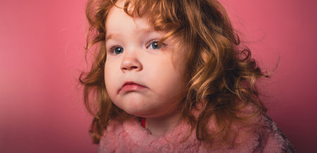 A young girl with curly, light hair sits in front of a plain pink background. She wears a pink blanket and gazes off into the distance, showing a thoughtful expression.の写真素材