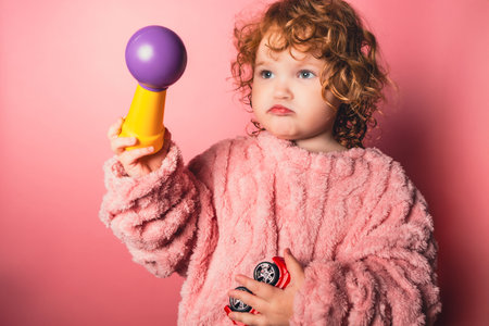 Child holds toys and looks thoughtful while standing against a pink background in a simple indoor...