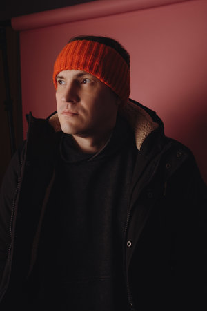 Man stands in front of a pink background wearing a black jacket and an orange headband indoors...