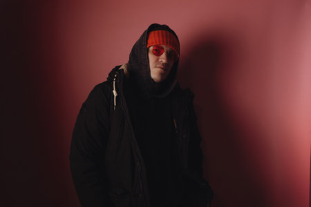 Man stands in a studio wearing a black jacket and hoodie with an orange beanie and sunglasses. He looks confidently at the camera against a pink backdrop.の写真素材