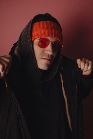 Man wears hoodie and orange hat with sunglasses in studio against pink background while posing for a...