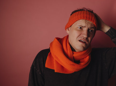 A man stands indoors wearing an orange scarf and headband, appearing confused while holding his...