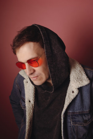 A man wears a hoodie with a jacket and red sunglasses. He looks down while standing against a plain...