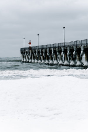 Winter pier extending over the sea, ice-covered pillars, street lamps, overcast sky, waves crashing at the shoreの写真素材