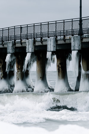 A winter pier with massive concrete pillars, covered in ice and icicles, extends over a choppy sea. Waves crash against the icy base under an overcast sky.の写真素材