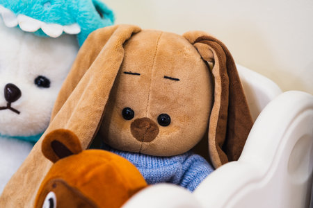 A plush brown bunny toy with long ears, wearing a blue knitted sweater, sits in a white container. Surrounded by other soft toys, creating a cozy sceneの写真素材