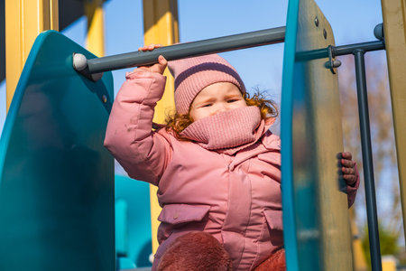 Little caucasian girl in a pink winter jumpsuit playing on a playground. Child enjoyment and...
