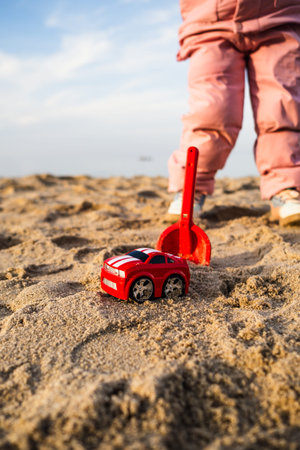 Red toy car and plastic shovel in the sand on the beach. Child play at the seaside. Concept of summer vacation and outdoor activity. Childhood leisure, travel and creative imaginationの写真素材