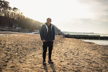 Man standing at the seaside during golden hour. Young guy wearing a denim jacket and beanie resting...