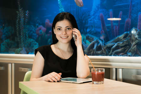 Stylish brunette woman sitting at a cafe drinking juice and talking on a cellphone in the background blue aquariumの写真素材