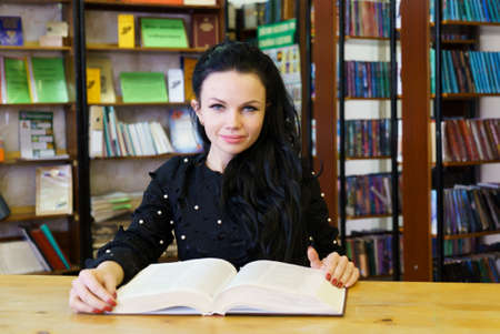 Attractive young woman sitting at table holding book posing for camera at libraryの写真素材