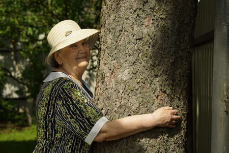 Elderly mature woman aged 80s dressed in white hat hugging tree trunk her hands in the forest. Woman is charged energy of nature. Relaxation in a summer dayの写真素材