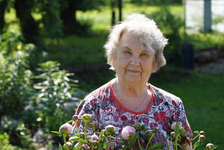 Portrait of old smiling woman in the park in the background of growing flowers as called peonies. Woman is charged energy of natureの写真素材