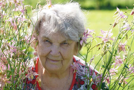 Portrait of old smiling woman in the park in the background of growing flowers as called peonies. Woman is charged energy of natureの写真素材