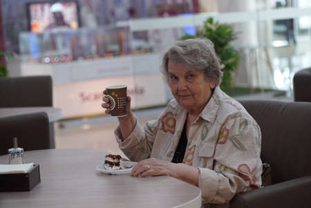 Portrait of old elderly smiling woman dressed in grey cotton shirt sitting in cafe holding cup of coffee. Wonderful moodの写真素材